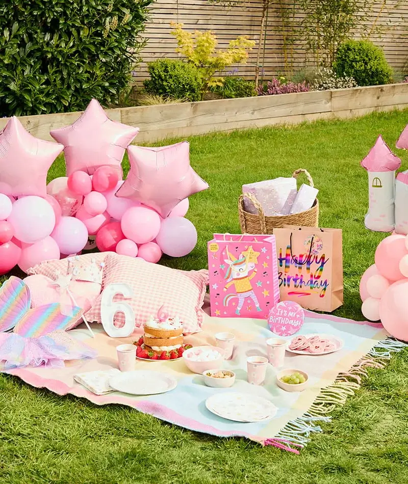 Pastel pink birthday picnic setup with balloons, gift bags, a birthday cake, party tableware, and a number 6 decoration on a picnic blanket. Pastel pink birthday picnic setup with balloons, gift bags, a birthday cake, party tableware, and a number 6 decoration on a picnic blanket.